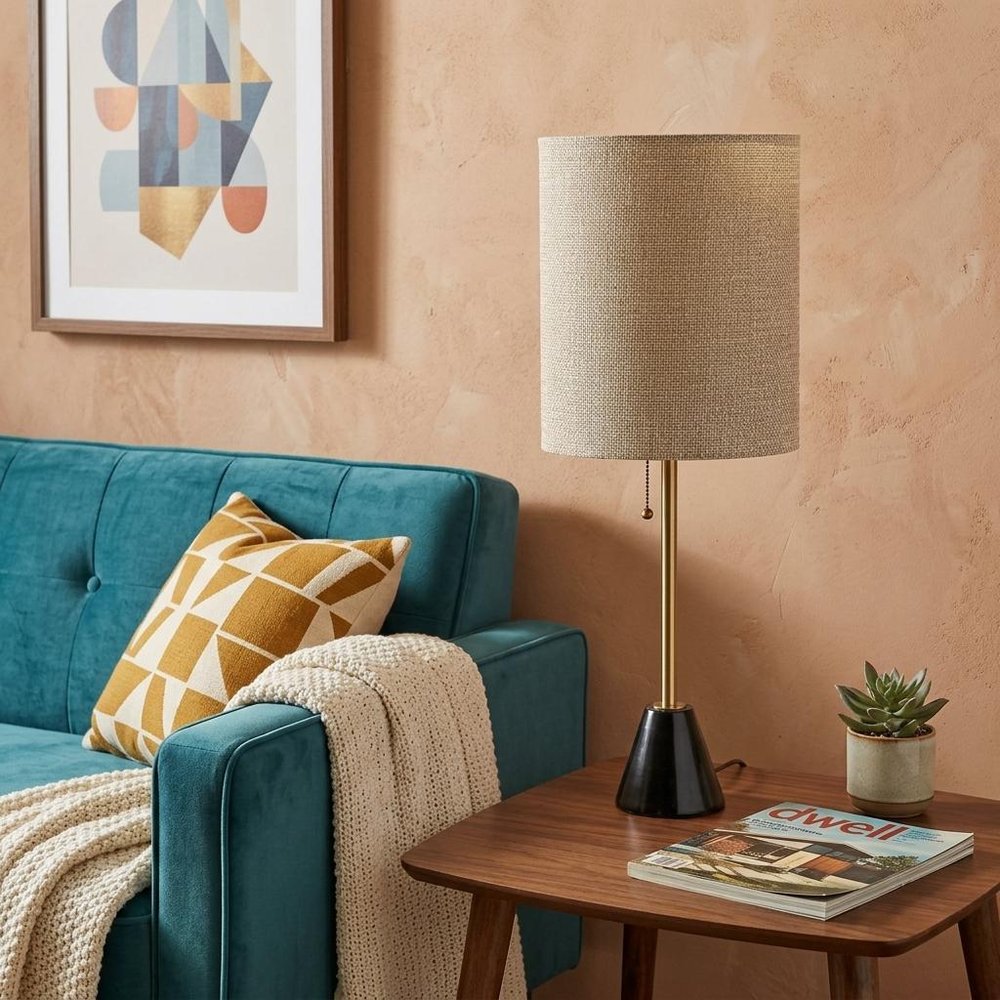 28" Black and Gold Marble and Metal Table Lamp With Beige Drum Shade displayed on a dark wood tabletop in a furnished interior against a tan wall.