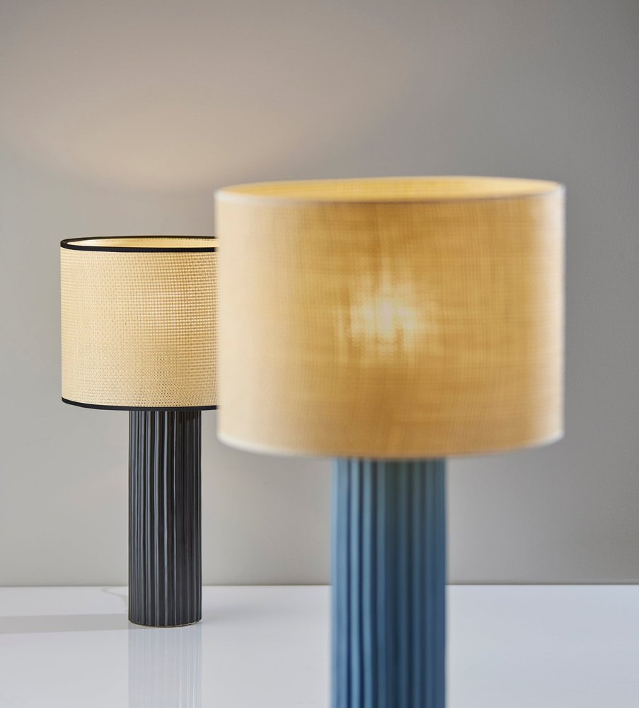 29" Black Ceramic Cylinder Table Lamp With Beige Drum Shade displayed on a tabletop in a furnished interior against a tan wall.