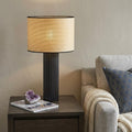 29" Black Ceramic Cylinder Table Lamp With Beige Drum Shade displayed on a dark wood tabletop in a furnished interior against a tan wall.