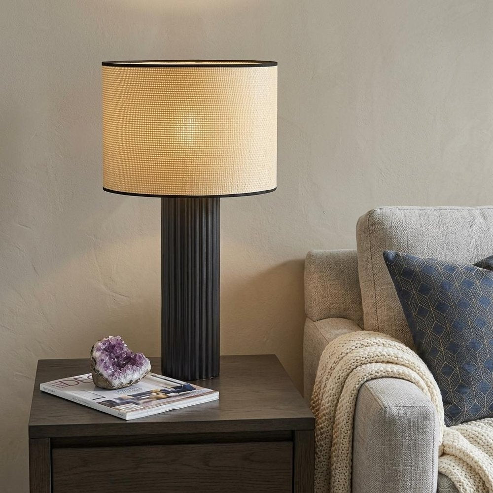 29" Black Ceramic Cylinder Table Lamp With Beige Drum Shade displayed on a dark wood tabletop in a furnished interior against a tan wall.