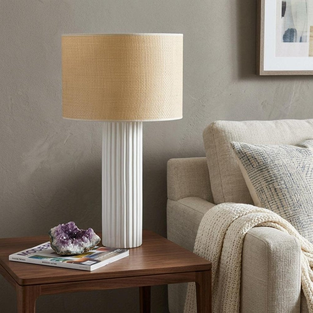 29" White Ceramic Cylinder Table Lamp With Beige Drum Shade displayed on a dark wood tabletop in a furnished interior against a gray wall.