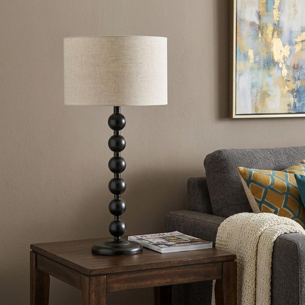 28" Black Solid Wood Candlestick Table Lamp With Off White Drum Shade displayed on a dark wood tabletop in a furnished interior against a tan wall.