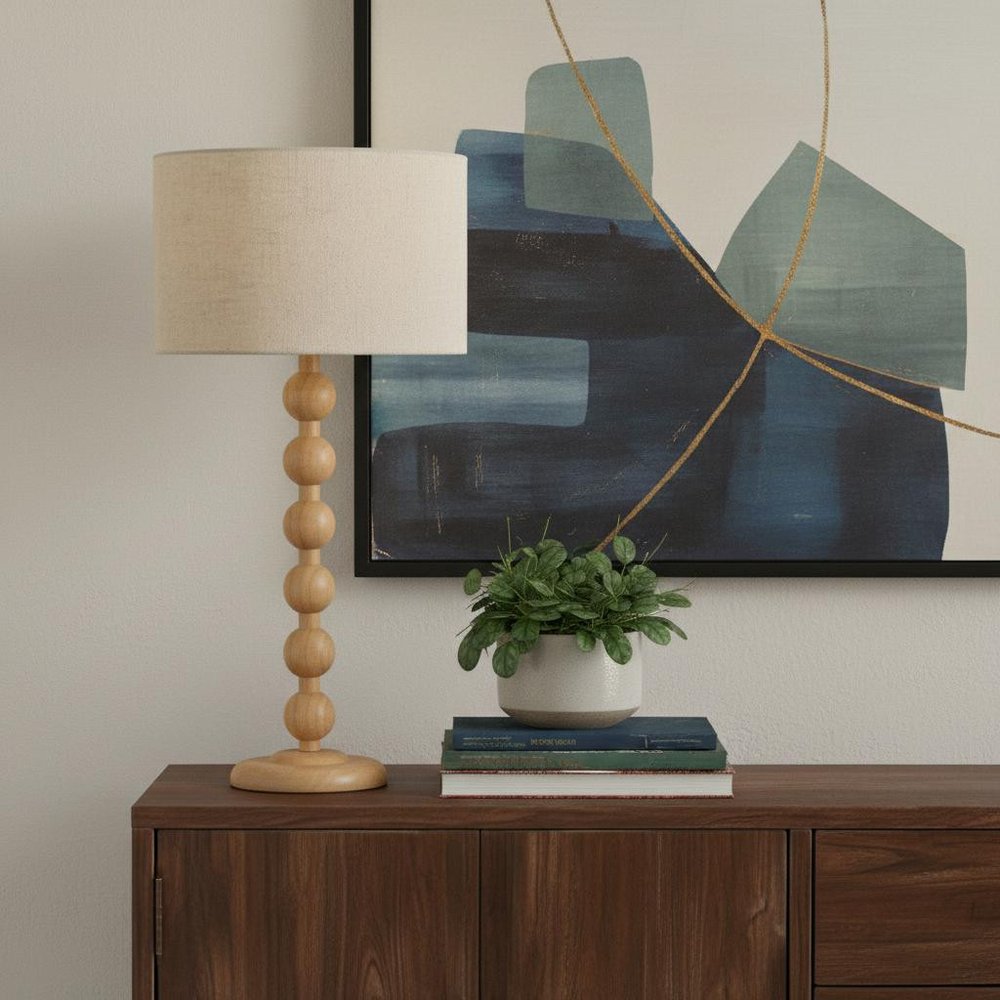 28" Brown Solid Wood Candlestick Table Lamp With Off White Drum Shade displayed on a dark wood tabletop in a furnished interior against a tan wall in off-white wood.