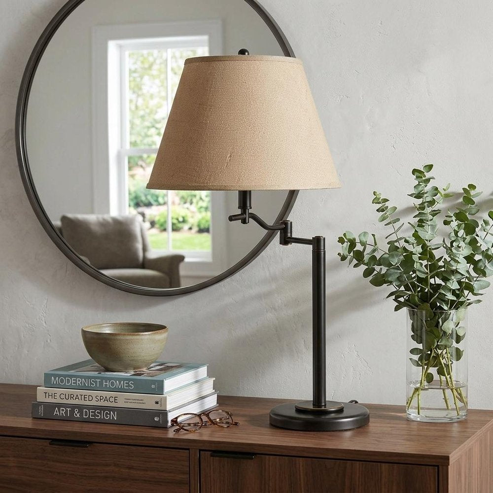 29" Bronze Metal Swing Arm Table Lamp With Off White Empire Shade displayed on a dark wood tabletop in a furnished interior against a light gray wall in off-white metal.