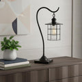 25" Bronze Metal Lantern Style Desk Lamp With Edison Bulb styled in a furnished interior with a light gray wall on a dark wood floor.