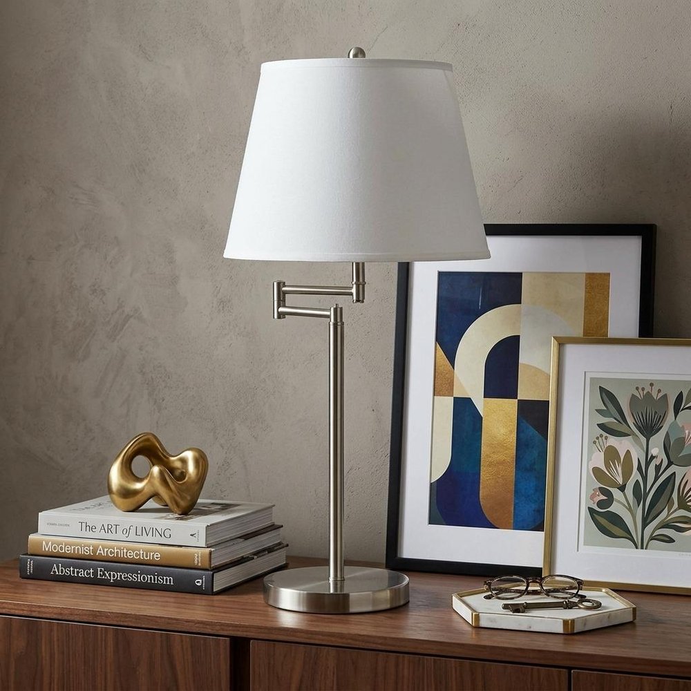28" Nickel Metal Table Lamp With Off White Empire Shade displayed on a dark wood tabletop in a furnished interior against a tan wall in silver metal.