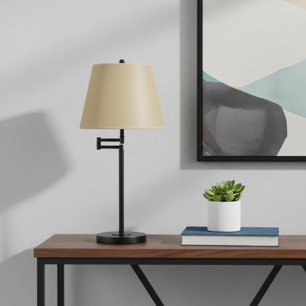 27" Bronze Metal Table Lamp With Tan Empire Shade displayed on a tabletop in a furnished interior against an off-white wall.