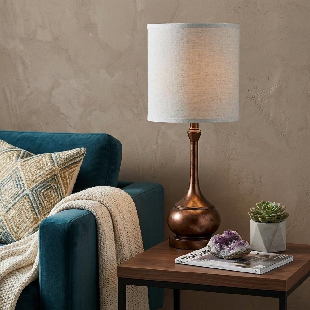 25" Rust Metal Round Table Lamp With White Drum Shade displayed on a dark wood tabletop in a furnished interior against a tan wall.