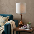 25" Rust Metal Round Table Lamp With White Drum Shade displayed on a dark wood tabletop in a furnished interior against a tan wall.