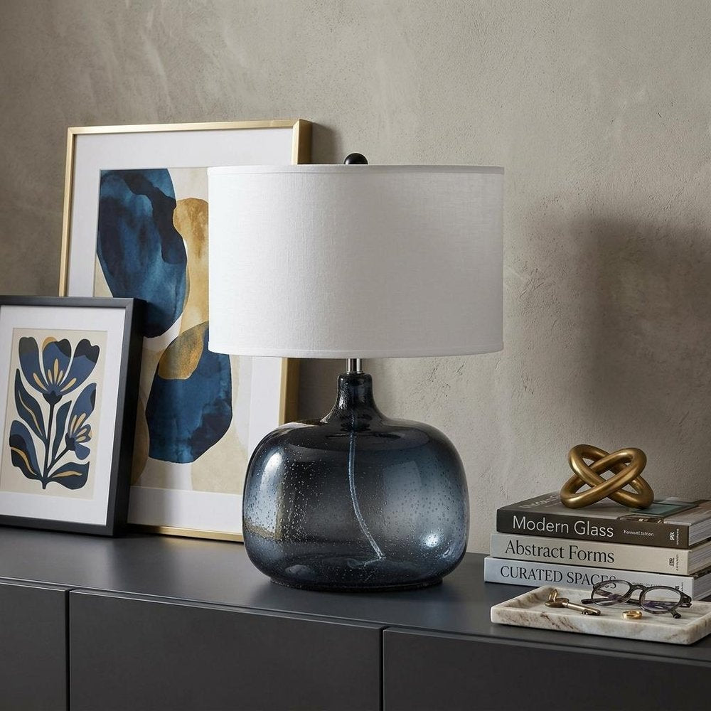 24" Dark Blue Glass Table Lamp With White Novelty Shade displayed on a tabletop in a furnished interior against a light gray wall.