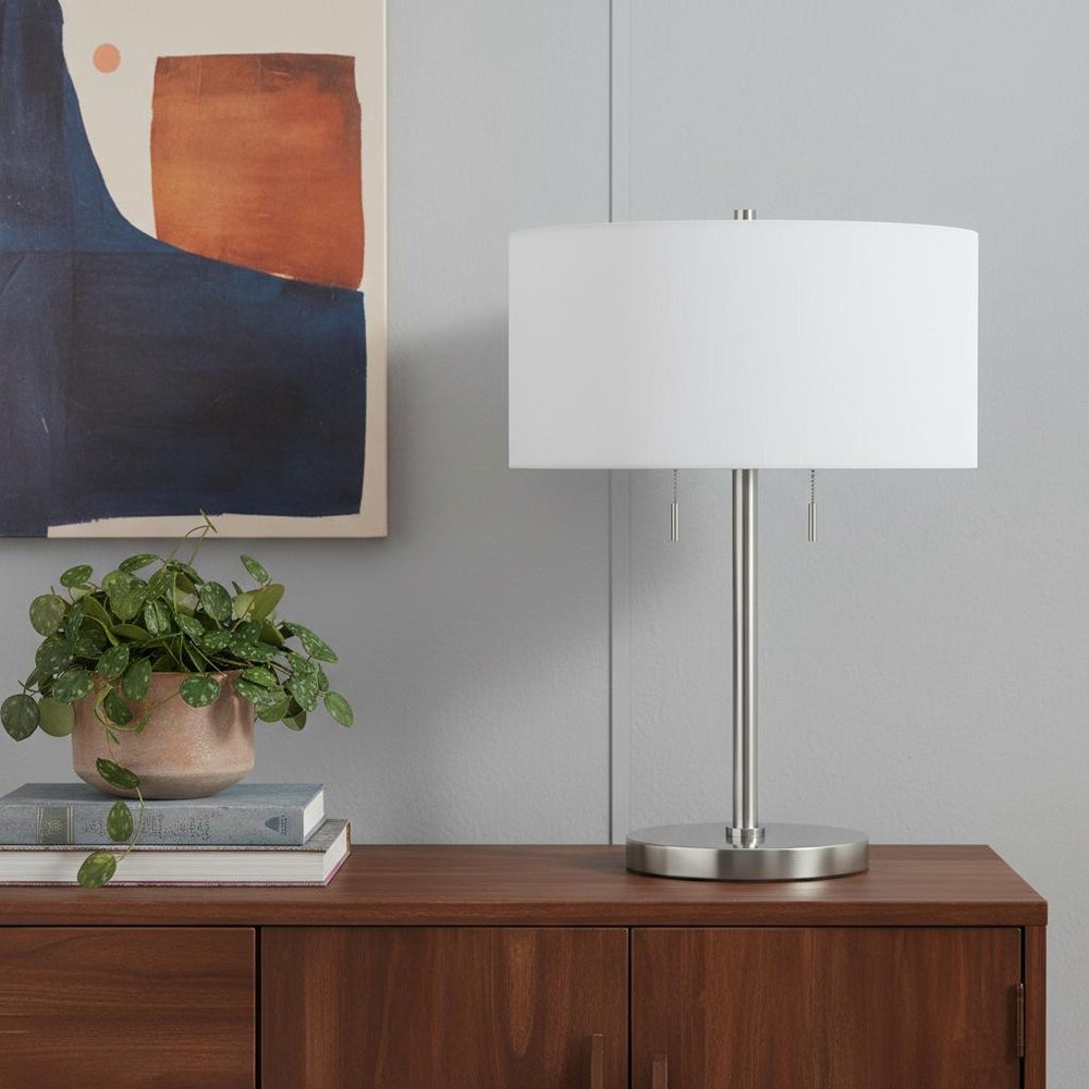 24" Nickel Metal Two Light Table Lamp With White Empire Shade displayed on a dark wood tabletop in a furnished interior against a light gray wall in silver metal.