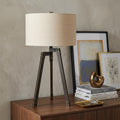 32" Charcoal Metal Tripod Table Lamp With Tan Drum Shade displayed on a dark wood tabletop in a furnished interior against a tan wall.