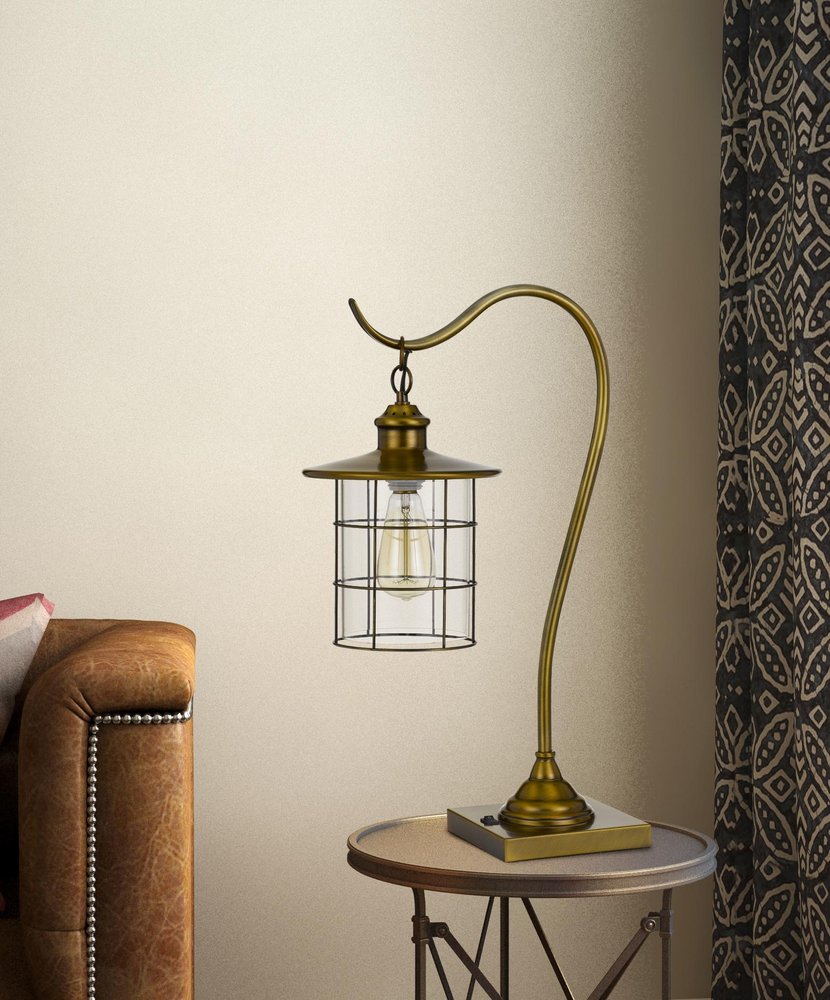 25" Antiqued Brass Metal Lantern Desk Lamp With Antiqued Brass Drum Shade styled in a furnished interior with a cream wall on a wood floor.