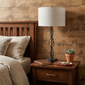 30" Black Metal Candlestick Table Lamp With Off White Drum Shade displayed on a dark wood tabletop in a furnished interior against a tan wall.