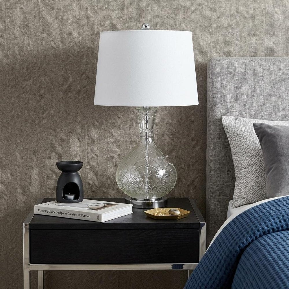 27" Clear Metal Round Table Lamp With White Shade displayed on a tabletop in a furnished interior against a light gray wall.