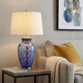 28" Blue Swirl Glass Table Lamp With White Empire Shade displayed on a dark wood tabletop in a furnished interior against a tan wall.