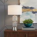 28" Nickel Metal Table Lamp With White Drum Shade displayed on a dark wood tabletop in a furnished interior against a light gray wall in silver metal.