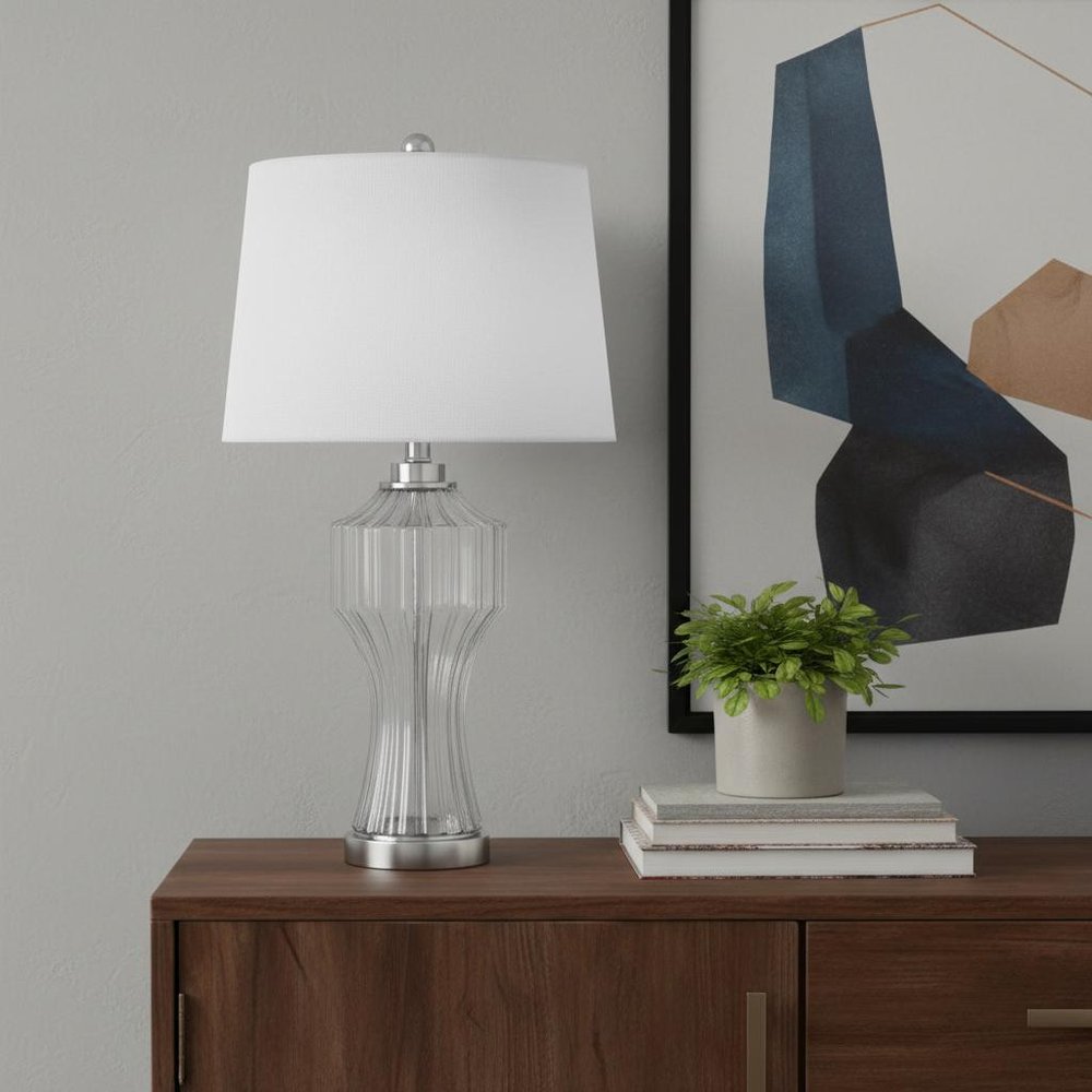26" Clear Glass Table Lamp With White Empire Shade displayed on a dark wood tabletop in a furnished interior against a light gray wall.