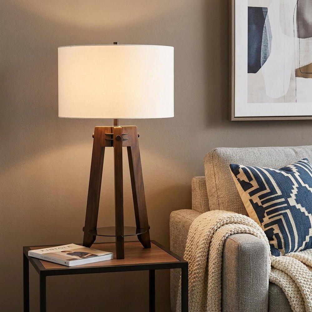 32" Brown Metal Tripod Table Lamp With Off White Drum Shade displayed on a dark wood tabletop in a furnished interior against a tan wall.