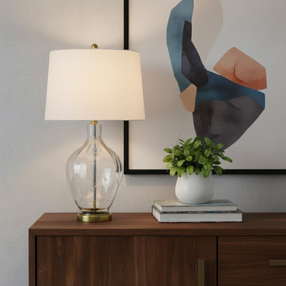 30" Clear Metal Table Lamp With Off White Drum Shade displayed on a dark wood tabletop in a furnished interior against a cream wall in off-white metal.