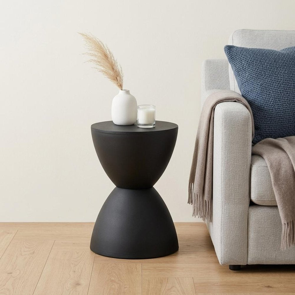 17" Black Heavy Duty Plastic Hourglass Outdoor Side Table styled in a furnished interior with an off-white wall on a wood floor.