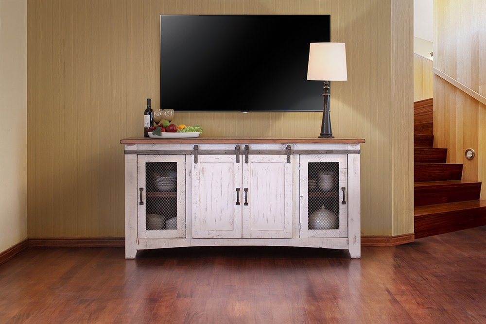 White solid wood TV stand with distressed finish and metal mesh doors, holding dishes and decor, in a room with a TV and stairs.