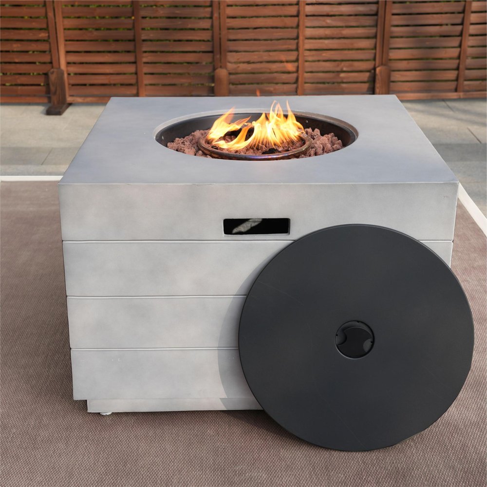 Gray aluminum square fire pit table with a black cover and flames, set on a patio with a wooden fence background.