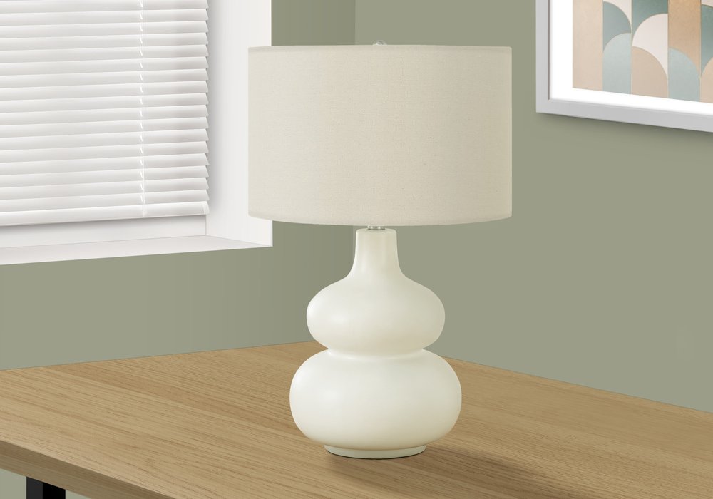 25" Cream Ceramic Gourd Table Lamp With Cream Drum Shade displayed on a wood tabletop in a furnished interior against an off-white wall.