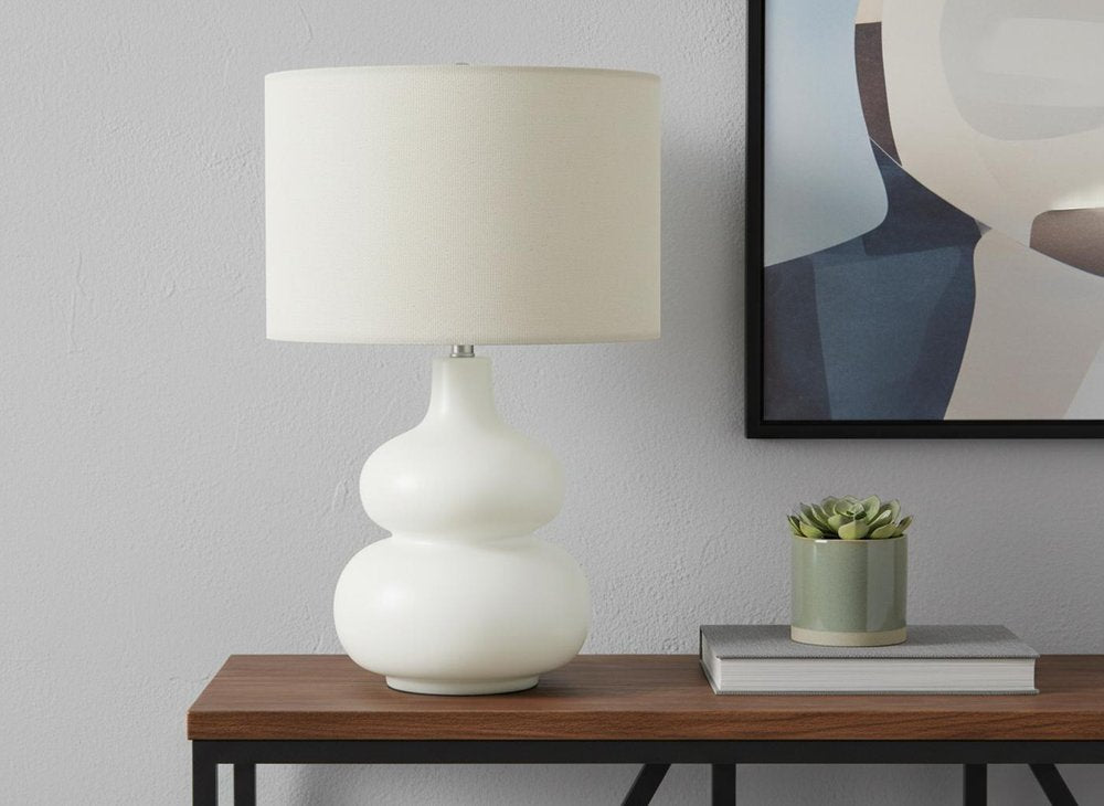 25" Cream Ceramic Gourd Table Lamp With Cream Drum Shade displayed on a dark wood tabletop in a furnished interior against a light gray wall.