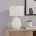 26" Cream Gourd Table Lamp With Cream Drum Shade displayed on a wood tabletop in a furnished interior against a light gray wall.