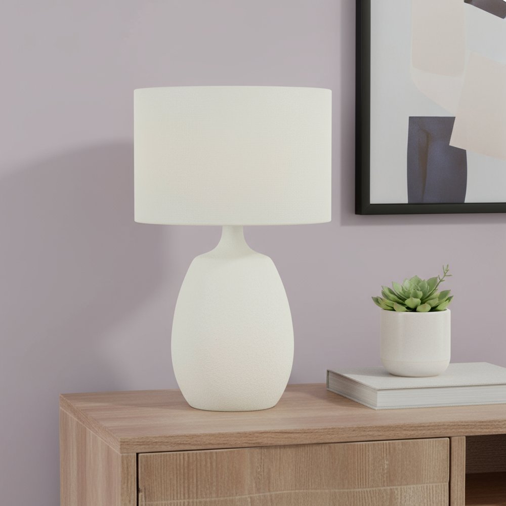 26" Cream Gourd Table Lamp With Cream Drum Shade displayed on a wood tabletop in a furnished interior against a light gray wall.