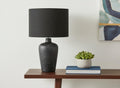 24" Black Ceramic Round Table Lamp With Black Drum Shade displayed on a tabletop in a furnished interior against an off-white wall.