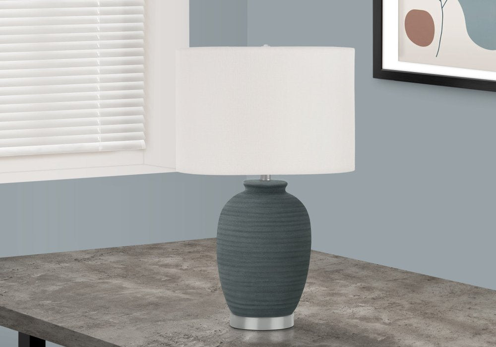 24" Blue and Silver Ceramic Round Table Lamp With Ivory Drum Shade displayed on a tabletop in a furnished interior against an off-white wall.