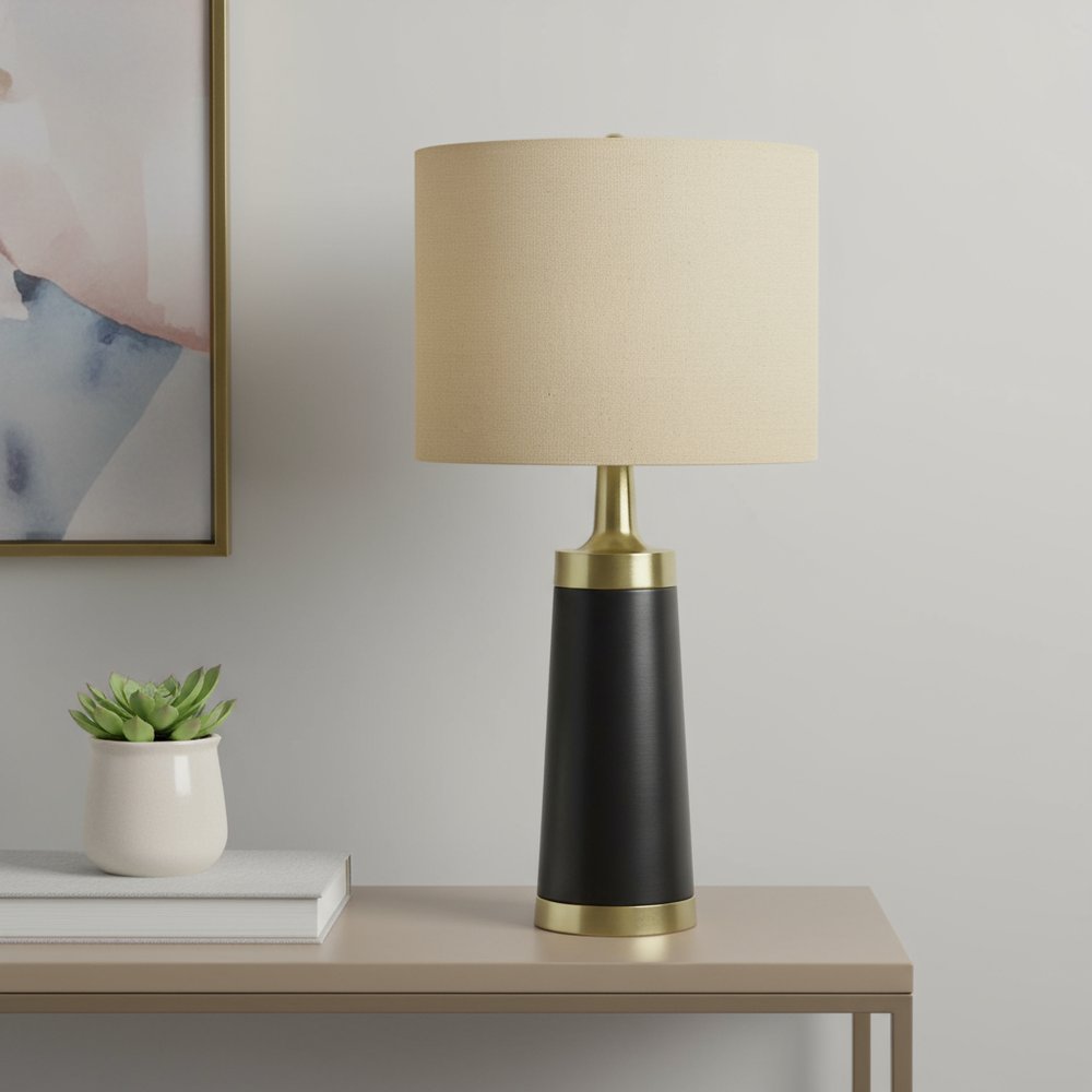 28" Black and Gold Metal Cylinder Table Lamp With Beige Drum Shade displayed on a tabletop in a furnished interior against a light gray wall.