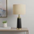 28" Black and Gold Metal Cylinder Table Lamp With Beige Drum Shade displayed on a tabletop in a furnished interior against a light gray wall.