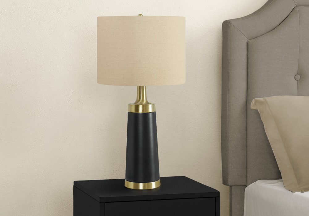 28" Black and Gold Metal Cylinder Table Lamp With Beige Drum Shade displayed on a tabletop in a furnished interior against a cream wall.