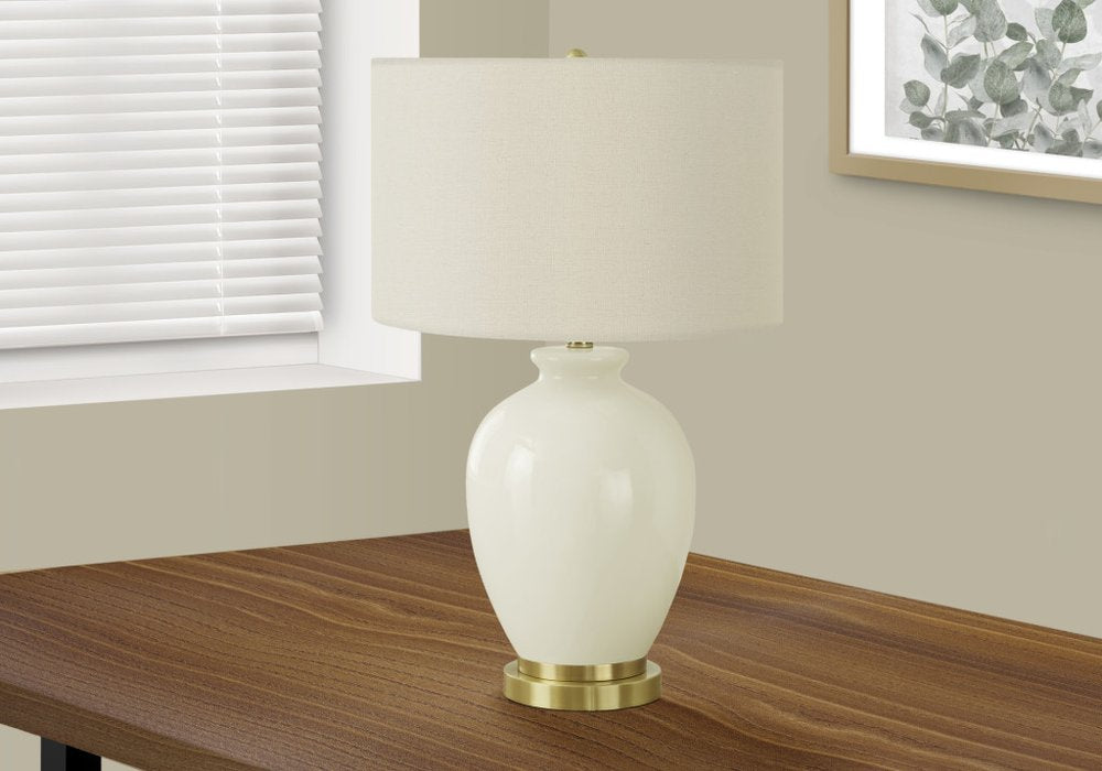 26" Gold and Ivory Ceramic Urn Table Lamp With Cream Drum Shade displayed on a dark wood tabletop in a furnished interior against an off-white wall.