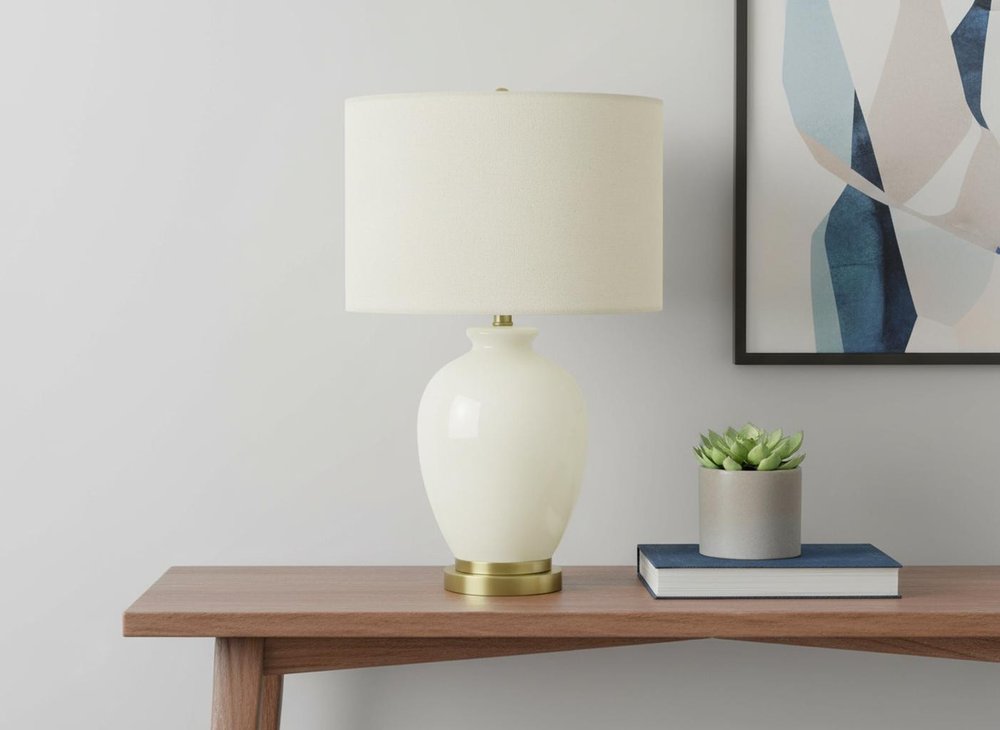 26" Gold and Ivory Ceramic Urn Table Lamp With Cream Drum Shade displayed on a tabletop in a furnished interior against an off-white wall.