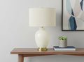 26" Gold and Ivory Ceramic Urn Table Lamp With Cream Drum Shade displayed on a tabletop in a furnished interior against an off-white wall.