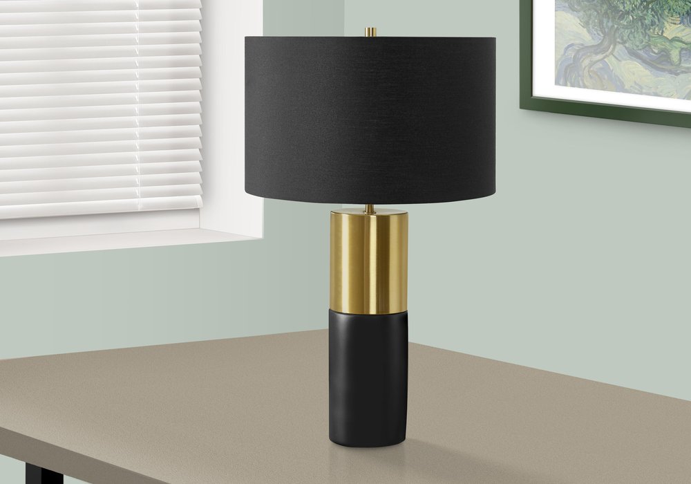 25" Black and Gold Concrete Cylinder Table Lamp With Black Drum Shade displayed on a wood tabletop in a furnished interior against an off-white wall.