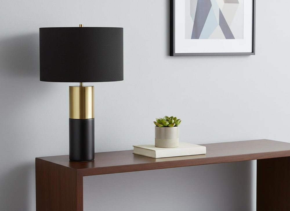 25" Black and Gold Concrete Cylinder Table Lamp With Black Drum Shade displayed on a tabletop in a furnished interior against an off-white wall.