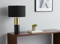 25" Black and Gold Concrete Cylinder Table Lamp With Black Drum Shade displayed on a tabletop in a furnished interior against an off-white wall.