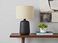 24" Black Ceramic Round Table Lamp With Beige Drum Shade displayed on a tabletop in a furnished interior against an off-white wall.