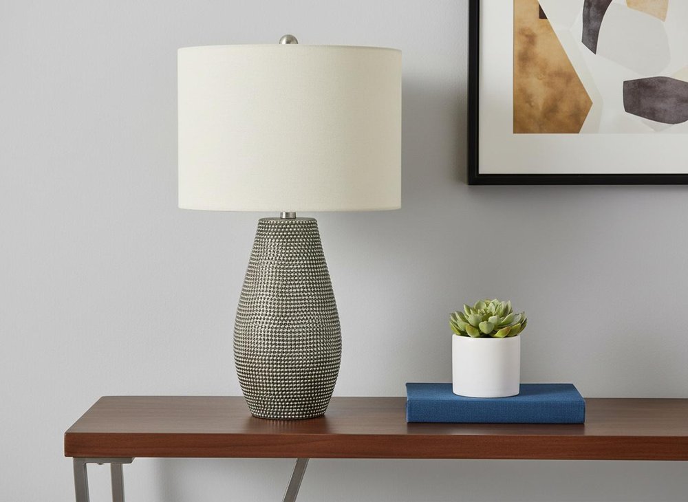 24" Gray Round Table Lamp With Ivory Drum Shade displayed on a dark wood tabletop in a furnished interior against a light gray wall.