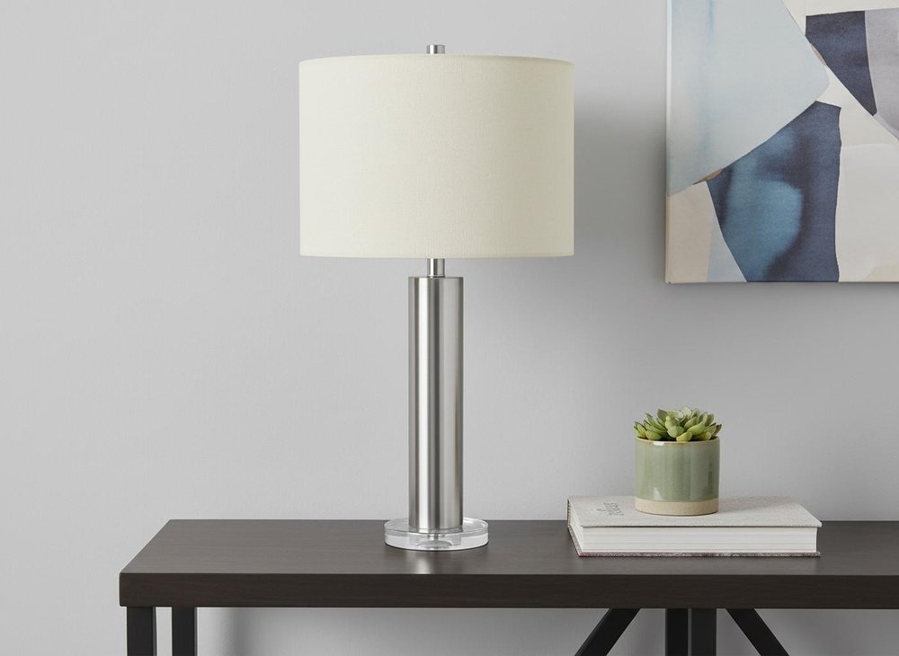 28" Silver Acrylic Cylinder Table Lamp With Ivory Drum Shade displayed on a tabletop in a furnished interior against a light gray wall.
