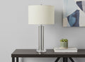 28" Silver Acrylic Cylinder Table Lamp With Ivory Drum Shade displayed on a tabletop in a furnished interior against a light gray wall.