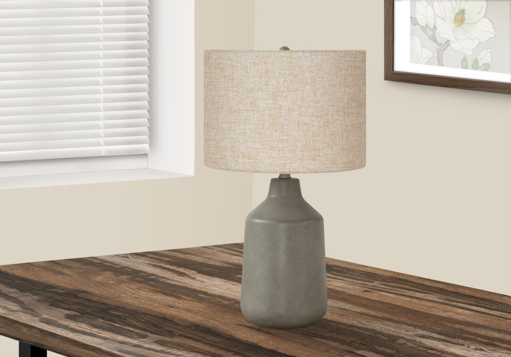 24" Gray Concrete Urn Table Lamp With Beige Drum Shade displayed on a dark wood tabletop in a furnished interior against an off-white wall.