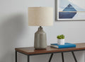 24" Gray Concrete Urn Table Lamp With Beige Drum Shade shown outdoors on a tiled patio.