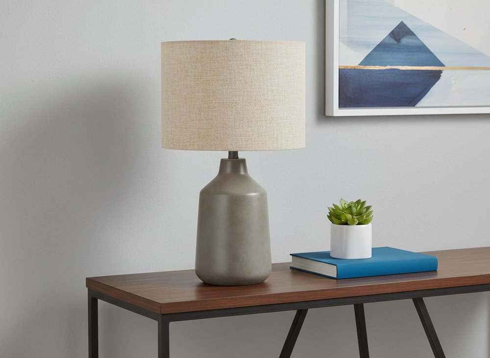 24" Gray Concrete Urn Table Lamp With Beige Drum Shade shown outdoors on a tiled patio.