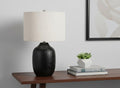 26" Black Ceramic Urn Table Lamp With Ivory Drum Shade displayed on a dark wood tabletop in a furnished interior against an off-white wall.
